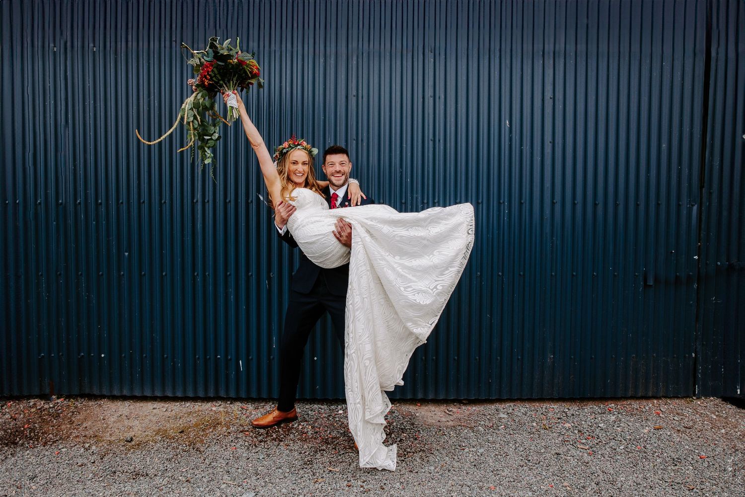 A wedding couple poses playfully in front of a teal corrugated metal wall with hanging greenery decorations.