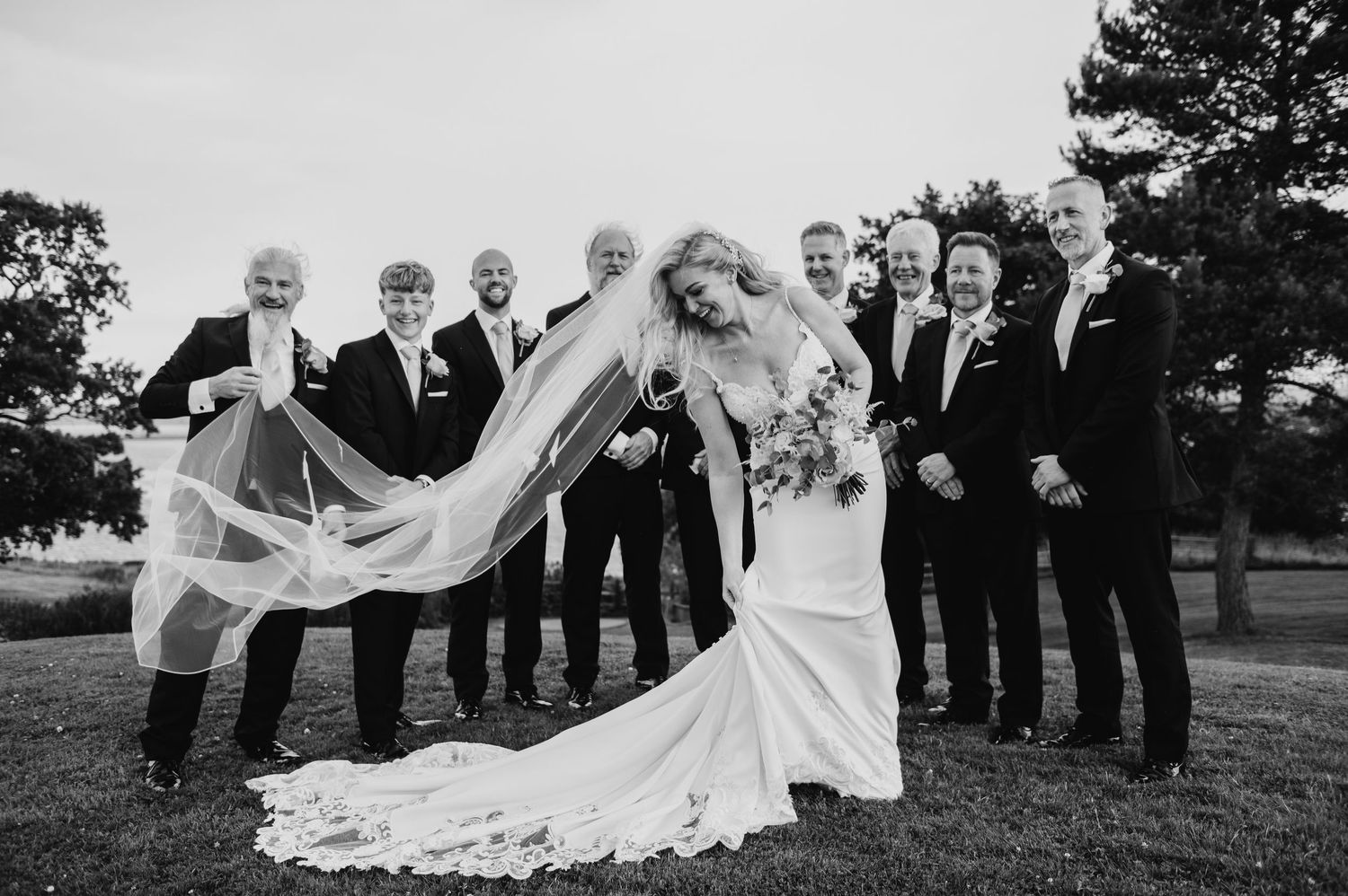A wedding party poses together on a lawn with the bride's veil flowing dramatically in the wind.