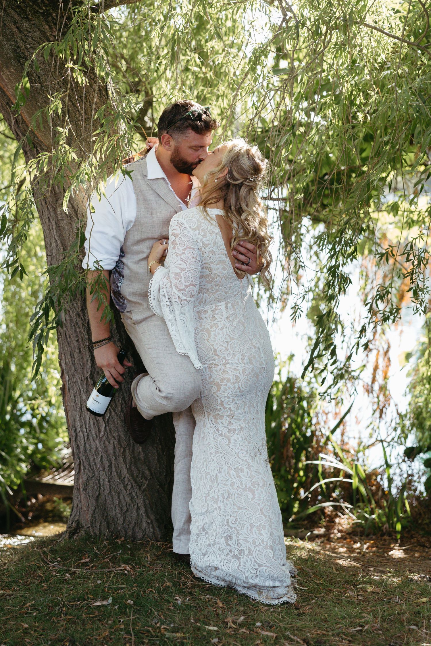 Romantic couple in white attire embraces beneath a weeping willow tree by the water on their wedding day.