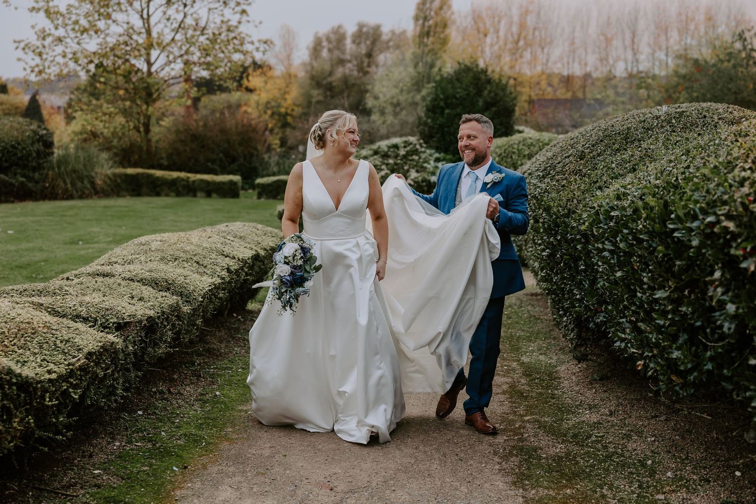 A couple walks together through a scenic garden pathway surrounded by lush hedges and trees on a sunny autumn day.