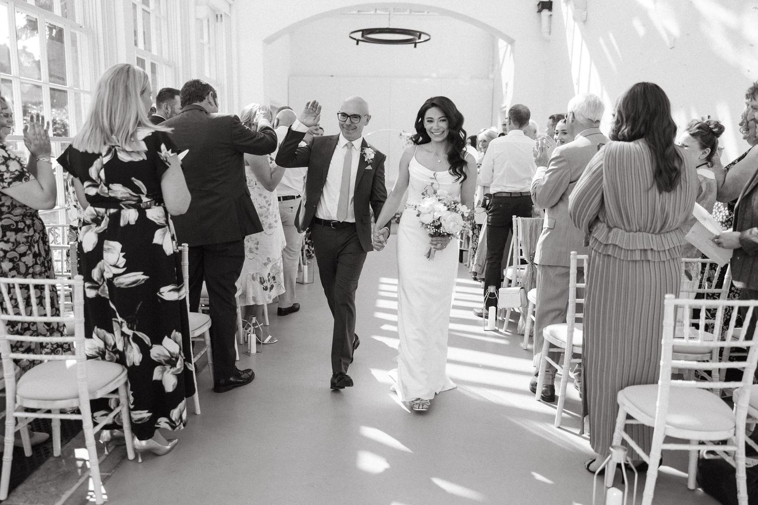 Wedding guests gather and mingle in an elegant white reception venue with sunlit windows and white chairs.