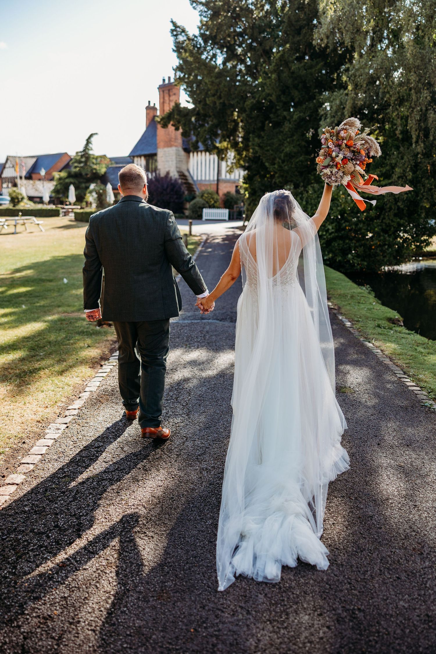 A couple walks down a gravel path on their wedding day with a beautiful estate building in the background.
