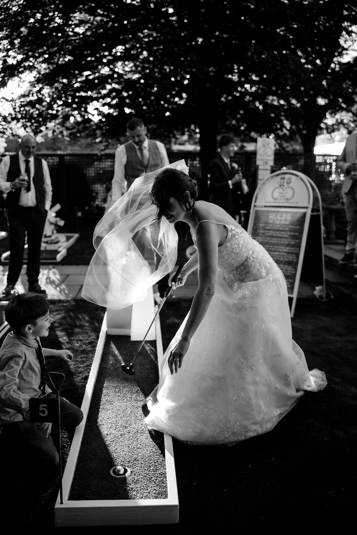 A person in a wedding dress plays miniature golf at an outdoor reception venue during a wedding celebration.
