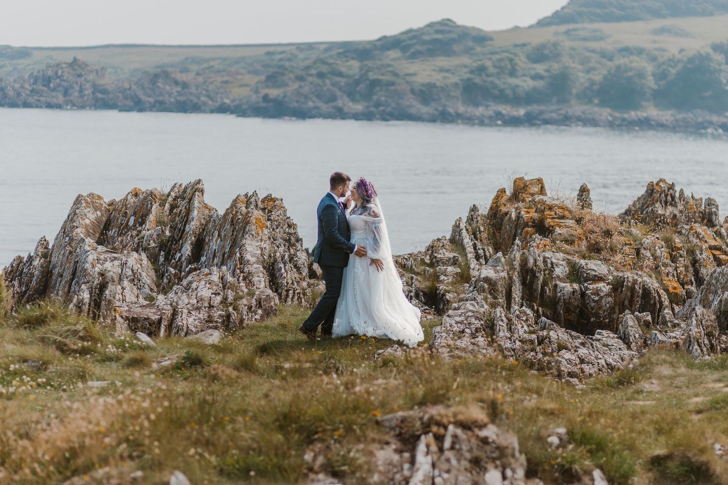 A romantic coastal wedding scene with a couple standing atop rugged cliffs overlooking a scenic loch in Scotland.