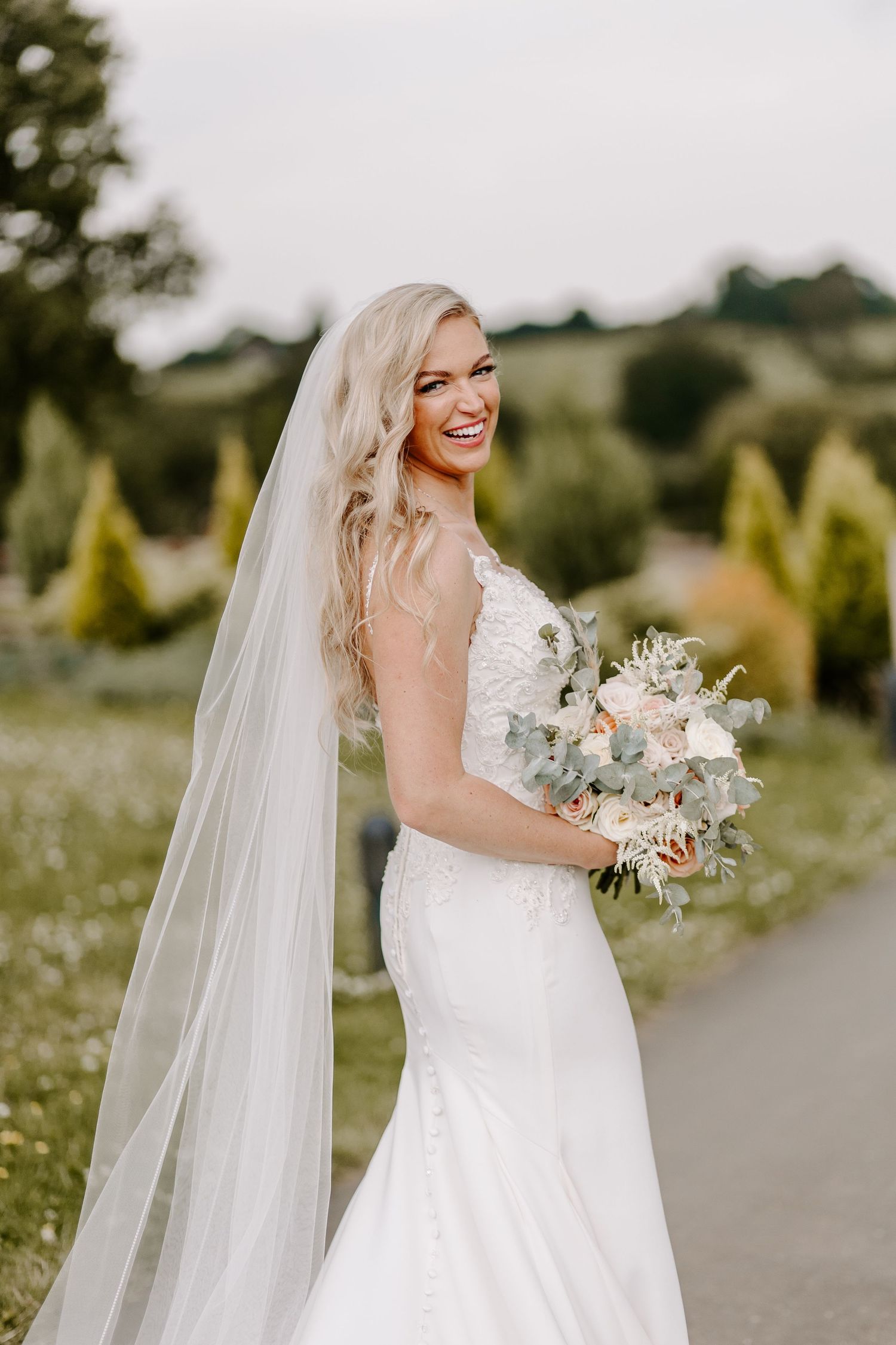 A bride in a fitted lace wedding gown holds a soft neutral bouquet while standing in a garden setting.