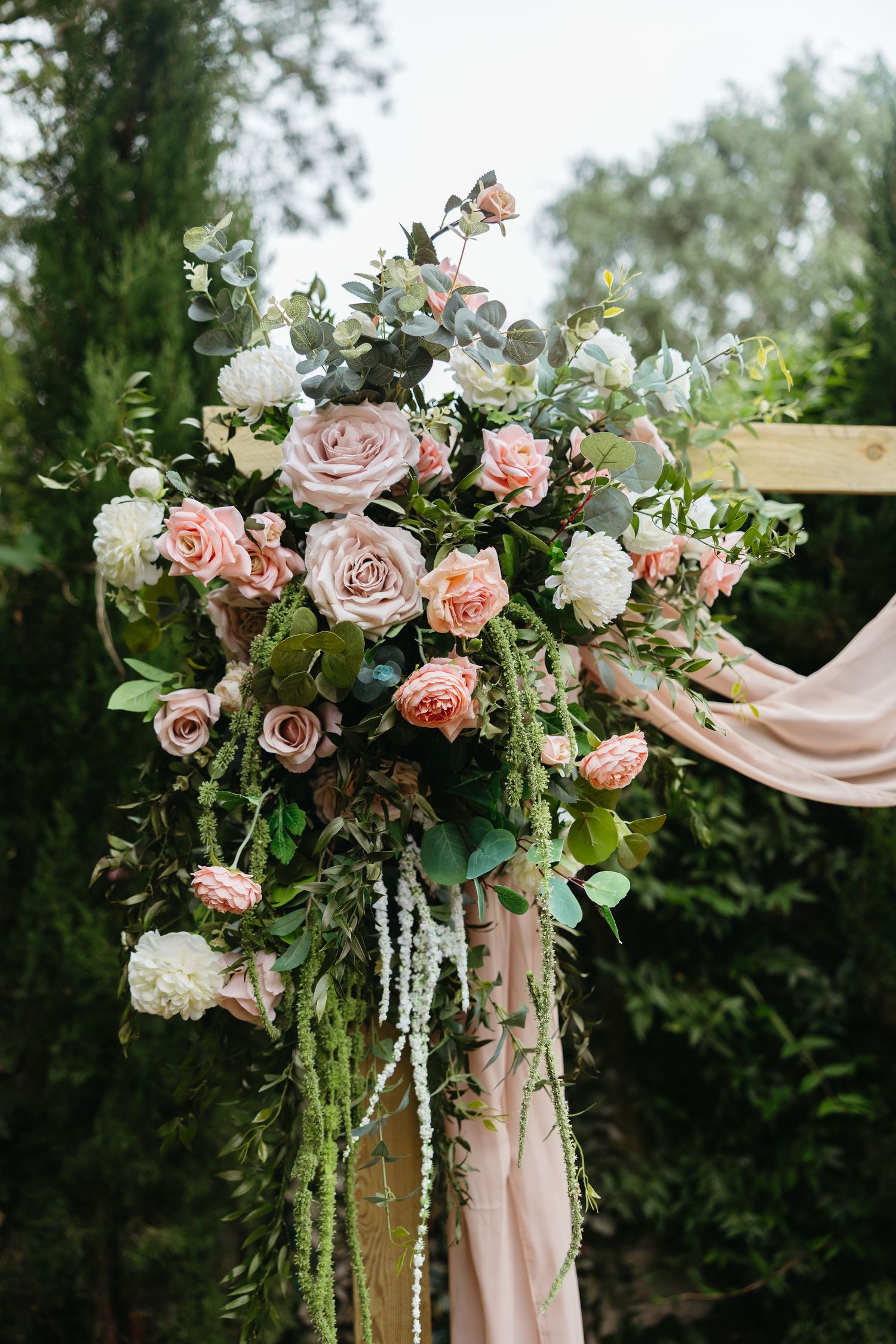 Elegant wedding arch adorned with blush pink roses, white flowers, and lush greenery with draped fabric outdoors.