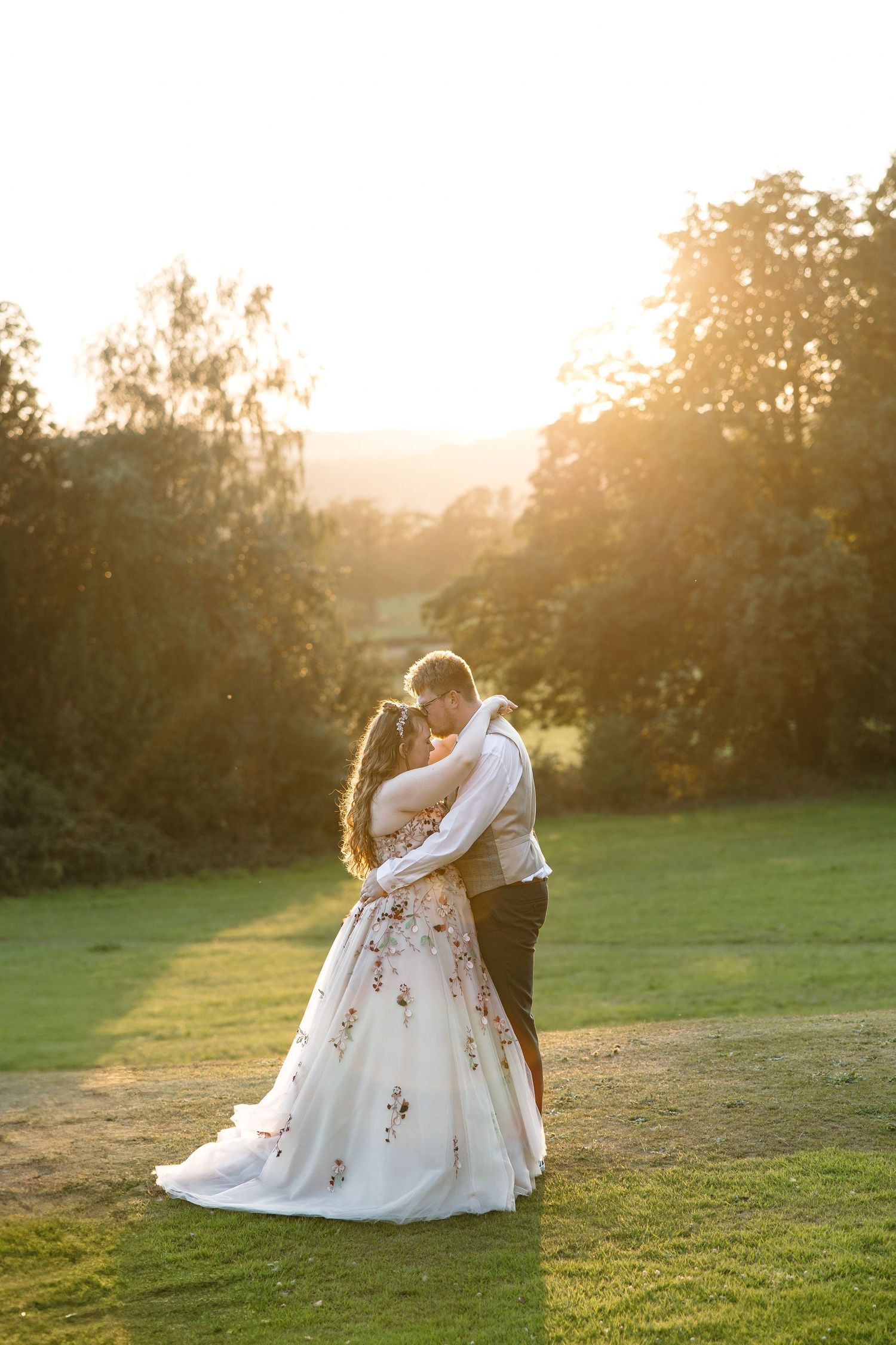Romantic couple embraces during sunset on a grassy hill with soft golden light filtering through trees.