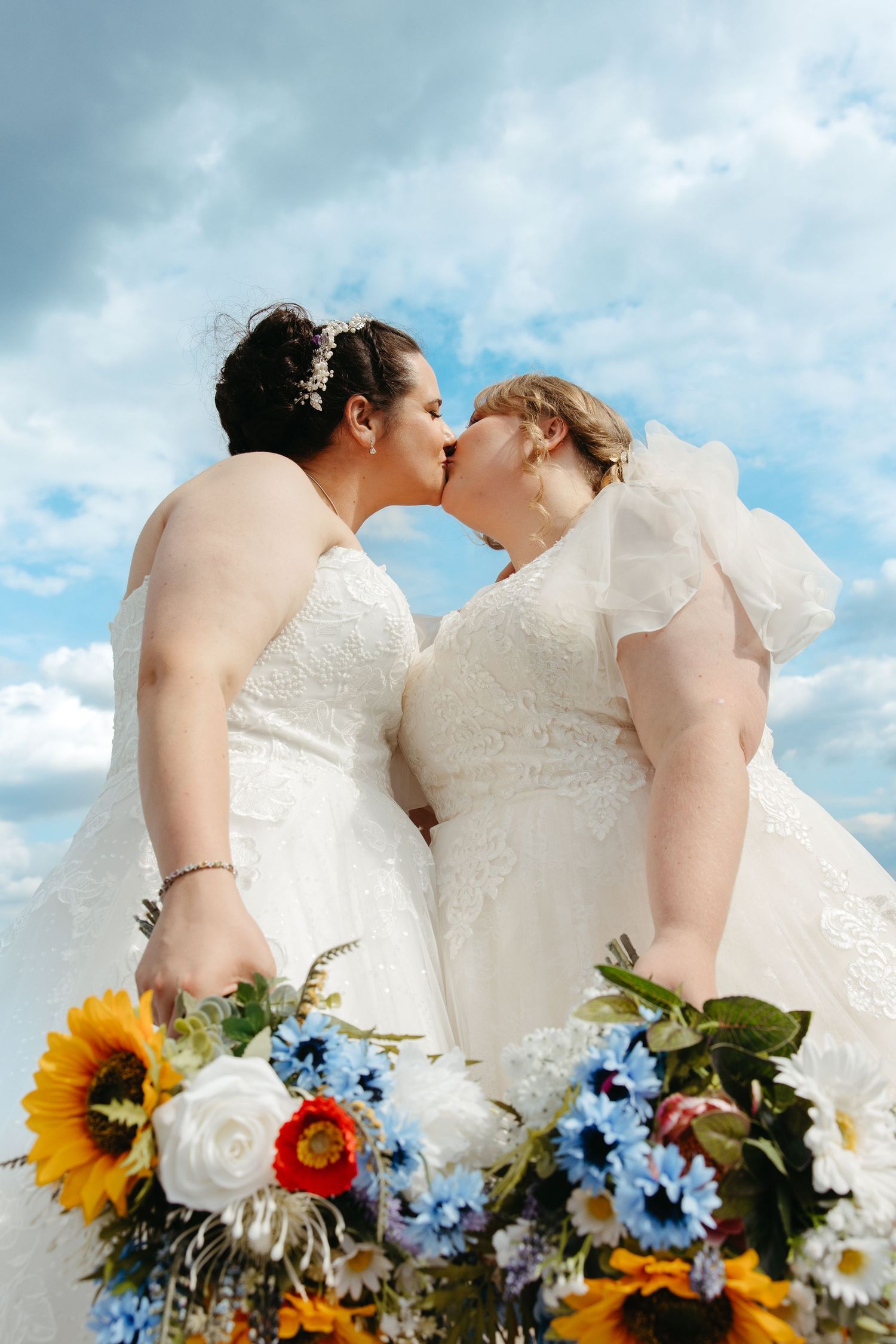 Two people in white wedding dresses share a romantic kiss against a blue sky with vibrant sunflowers and blue daisies.