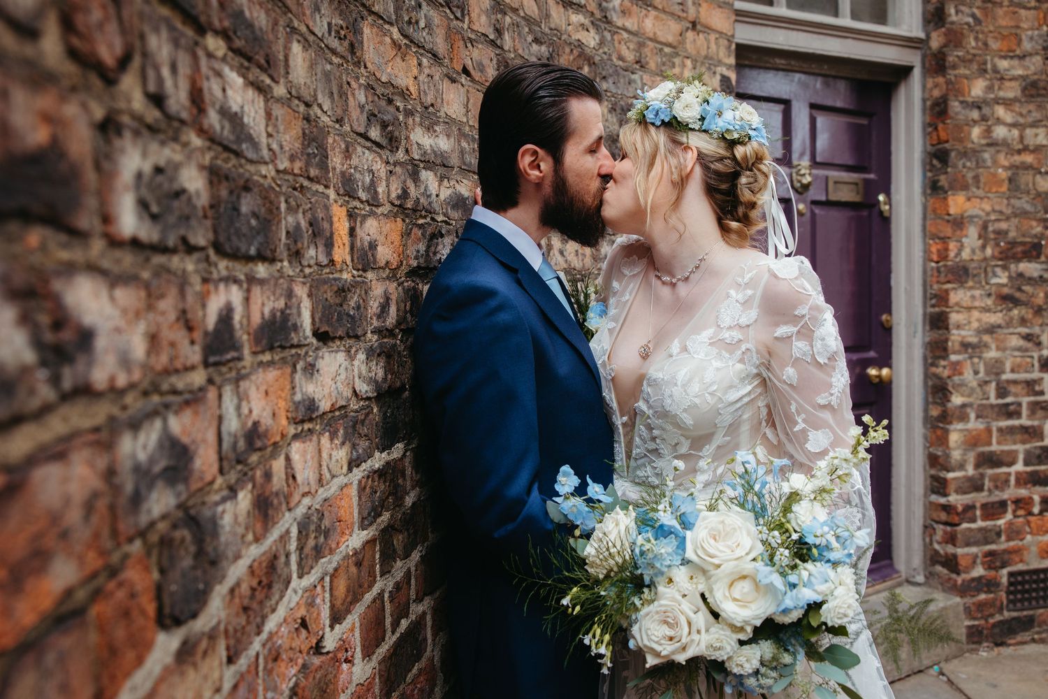 Romantic wedding couple embracing against a rustic brick wall with soft natural lighting.