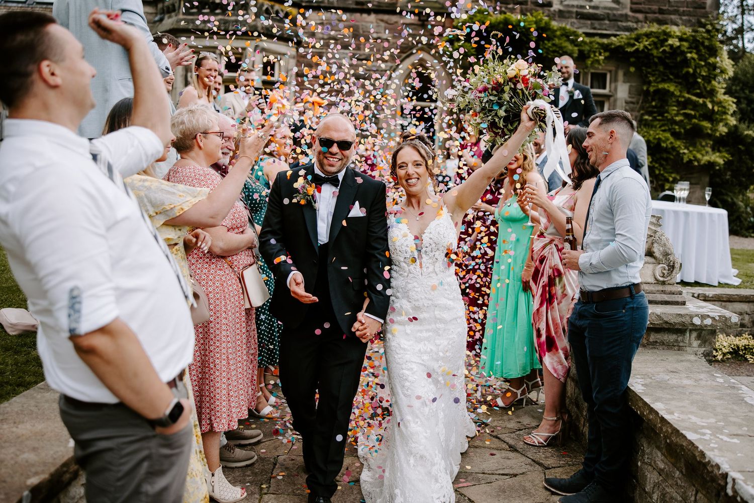 Wedding guests shower newlyweds with pink and white flower confetti during outdoor ceremony celebration.