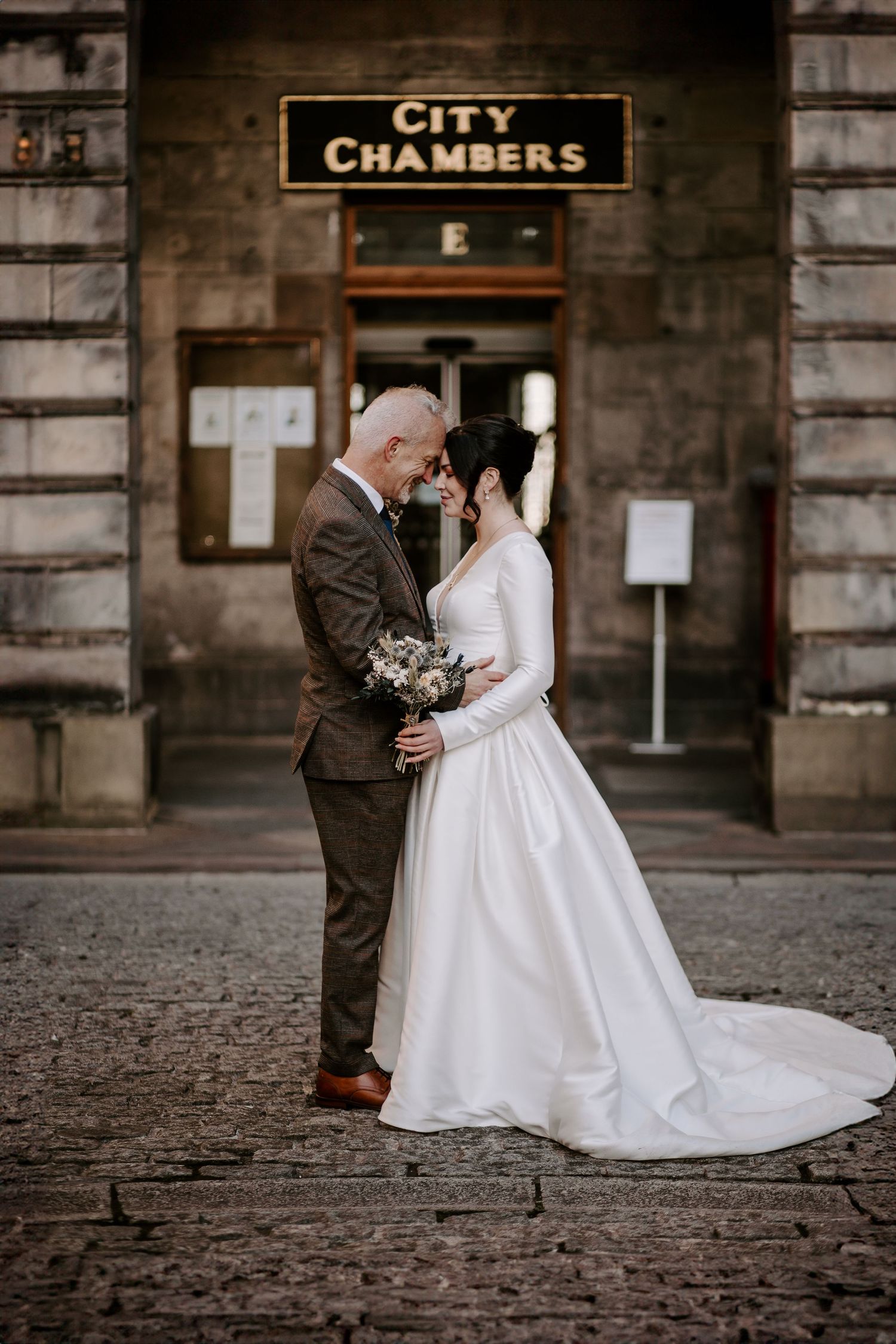 A romantic wedding photo of a bride and groom embracing outside City Chambers building on cobblestone street.