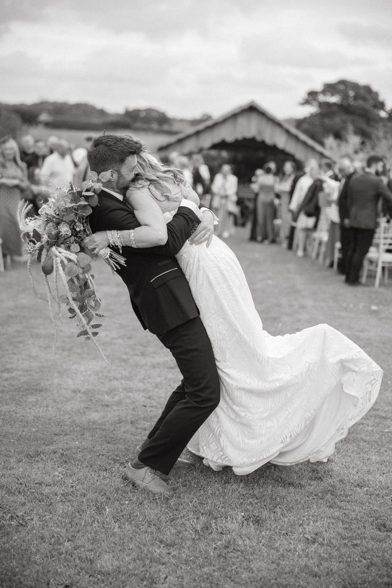 A romantic black and white wedding photo with a dip during a first dance on a lawn with guests looking on.