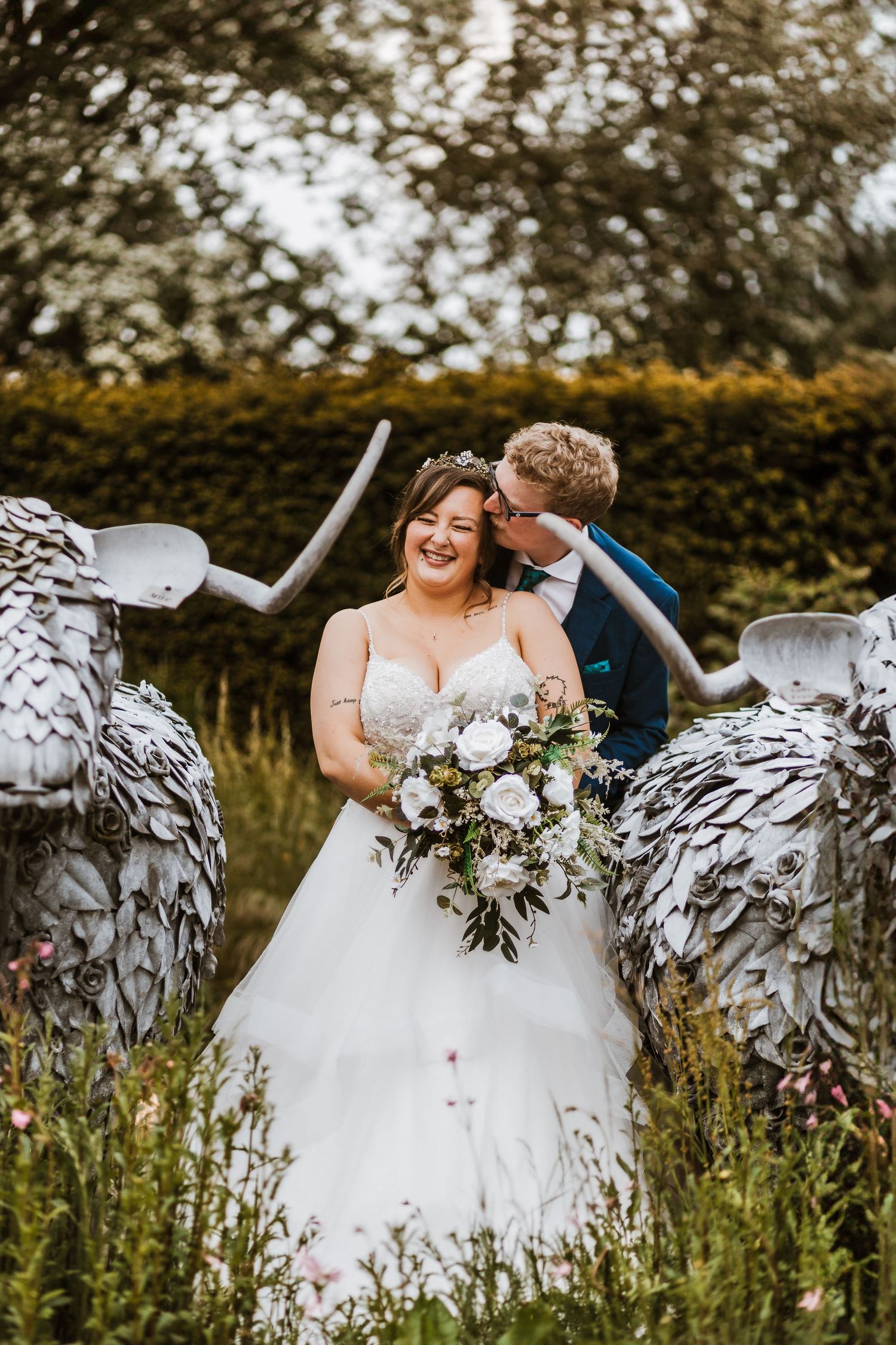 A wedding couple shares a joyful moment between two large metal swan sculptures in a garden setting.