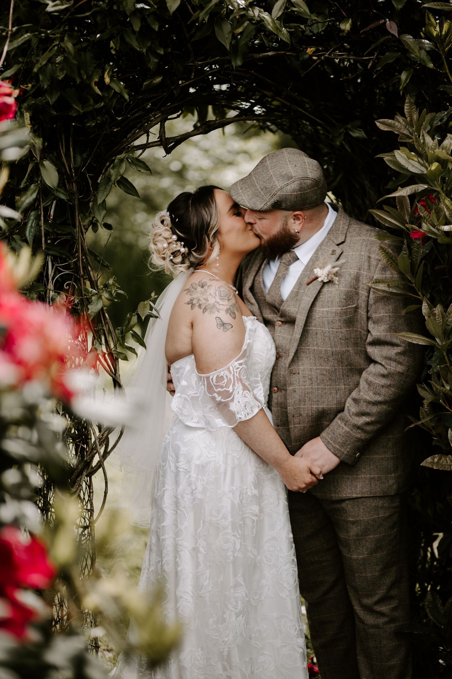 A romantic wedding moment captured in a garden archway surrounded by lush greenery and pink flowers.
