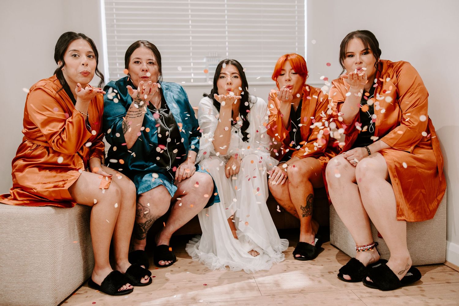 Wedding party in orange silk robes blowing confetti and celebrating together on a couch.