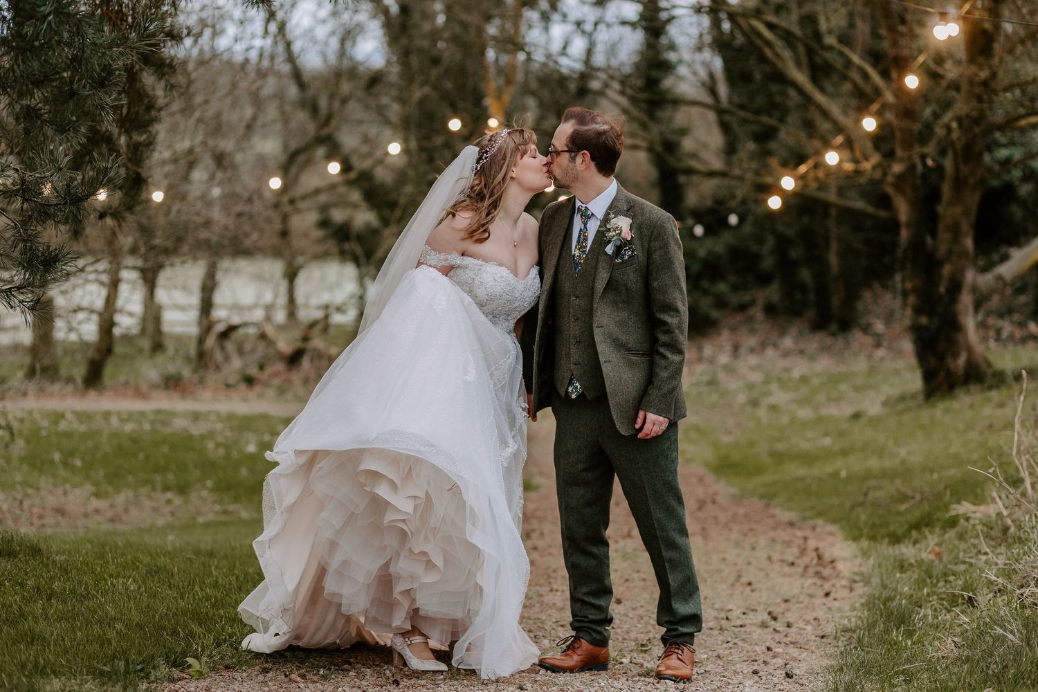 A couple embrace on a rustic path under string lights during their outdoor autumn wedding ceremony.