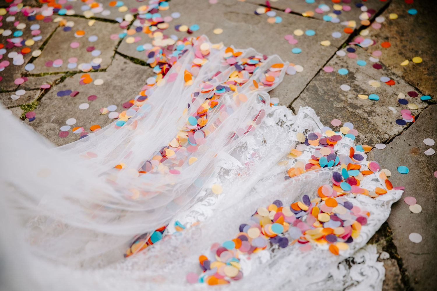 Colorful paper confetti scattered across a white wedding dress train and stone ground during a celebration.