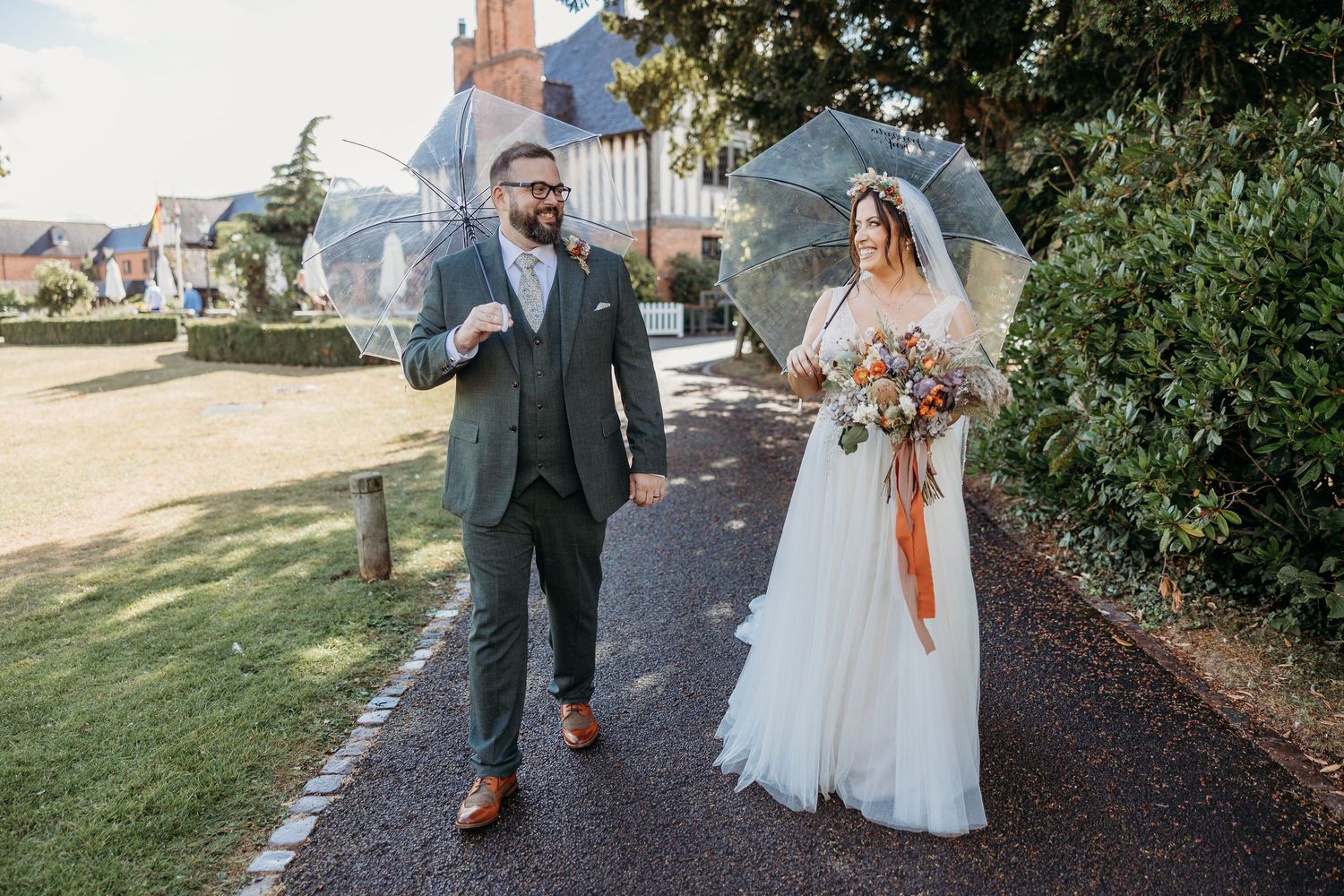 A couple walks along a garden path in formal wedding attire with the bride holding a vibrant autumn bouquet.