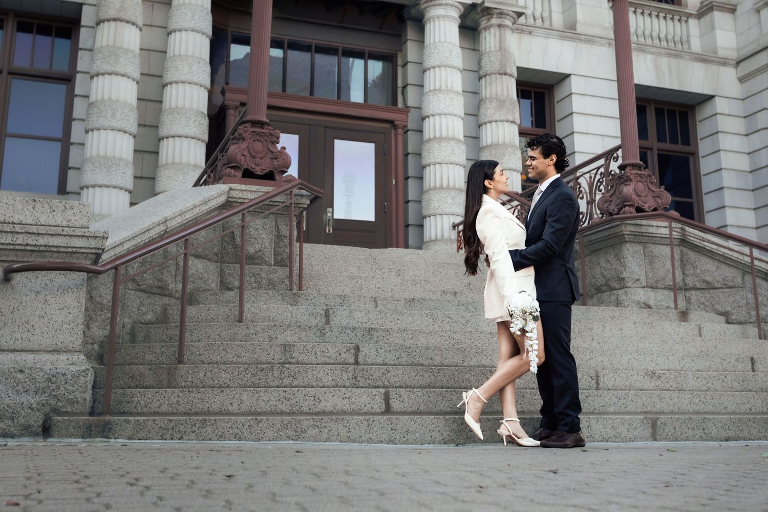 Couple in formal attire standing on stone steps outside a grand building, sharing a romantic moment.