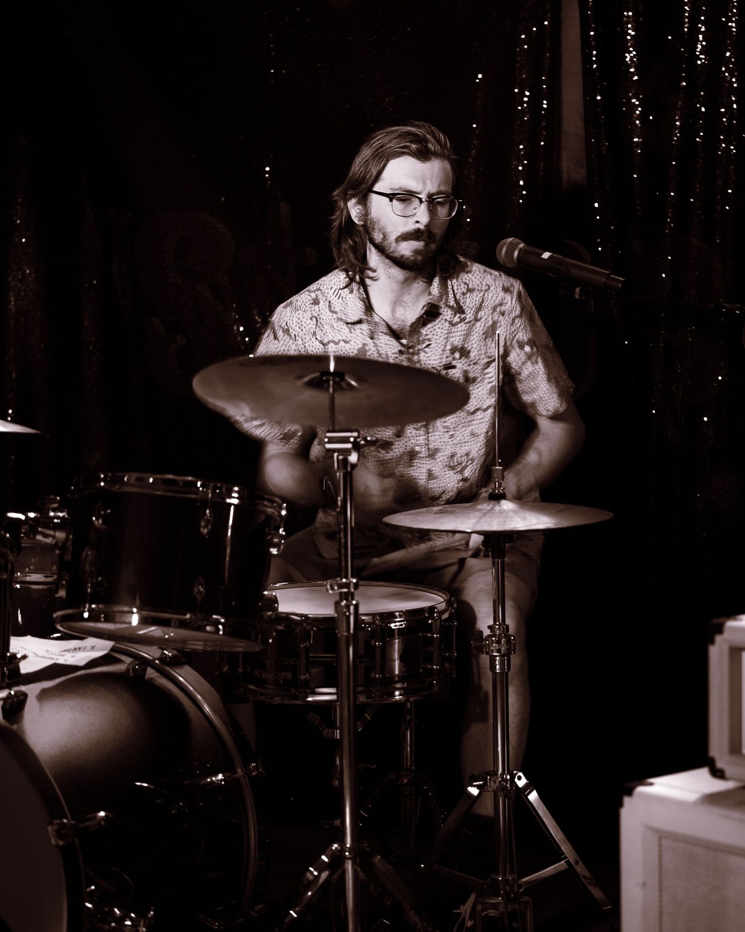 Bearded drummer with glasses playing a drum kit on a dimly lit stage.