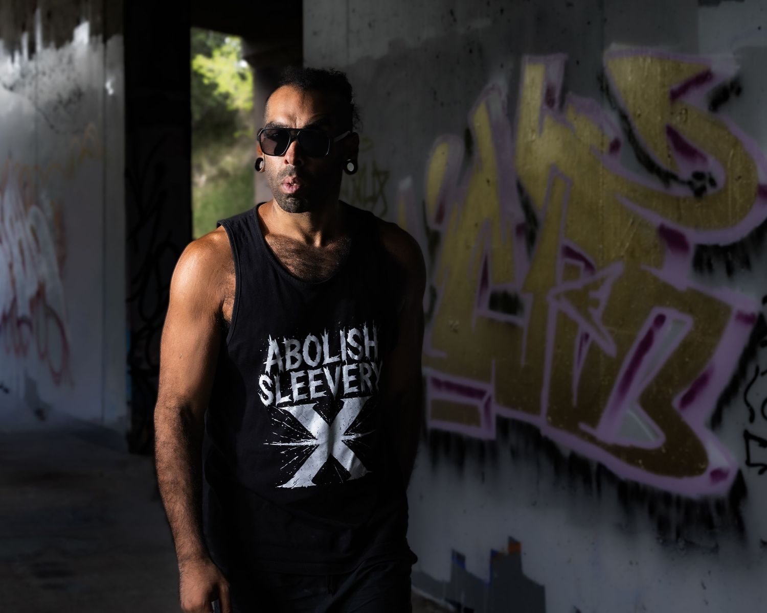 A person wearing a black sleeveless shirt poses against graffiti-covered walls in an urban setting.