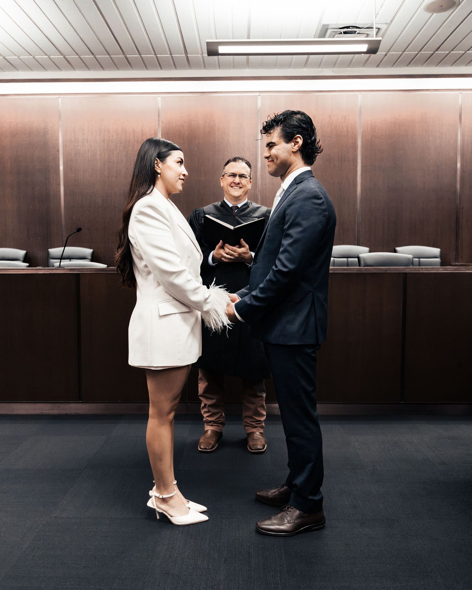 Couple exchanging vows in a courthouse ceremony with an officiant holding a book in a wood-paneled courtroom.