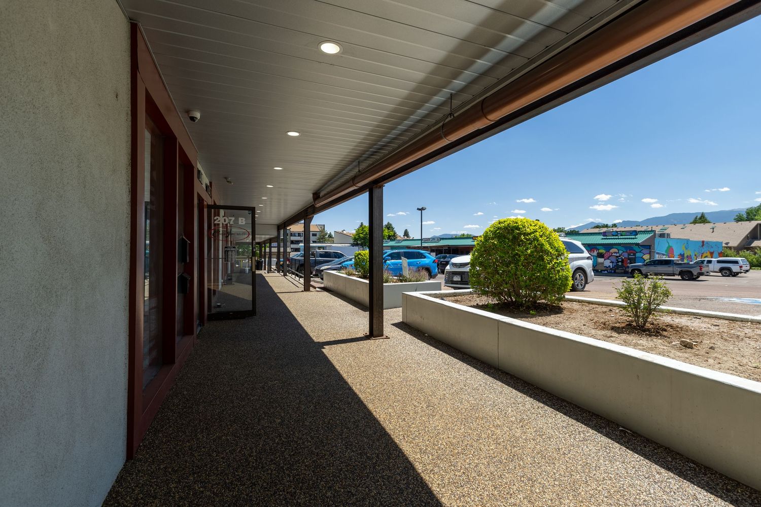 Covered walkway outside a motel with parked cars, blue sky, and a neatly trimmed green shrub in a garden bed.