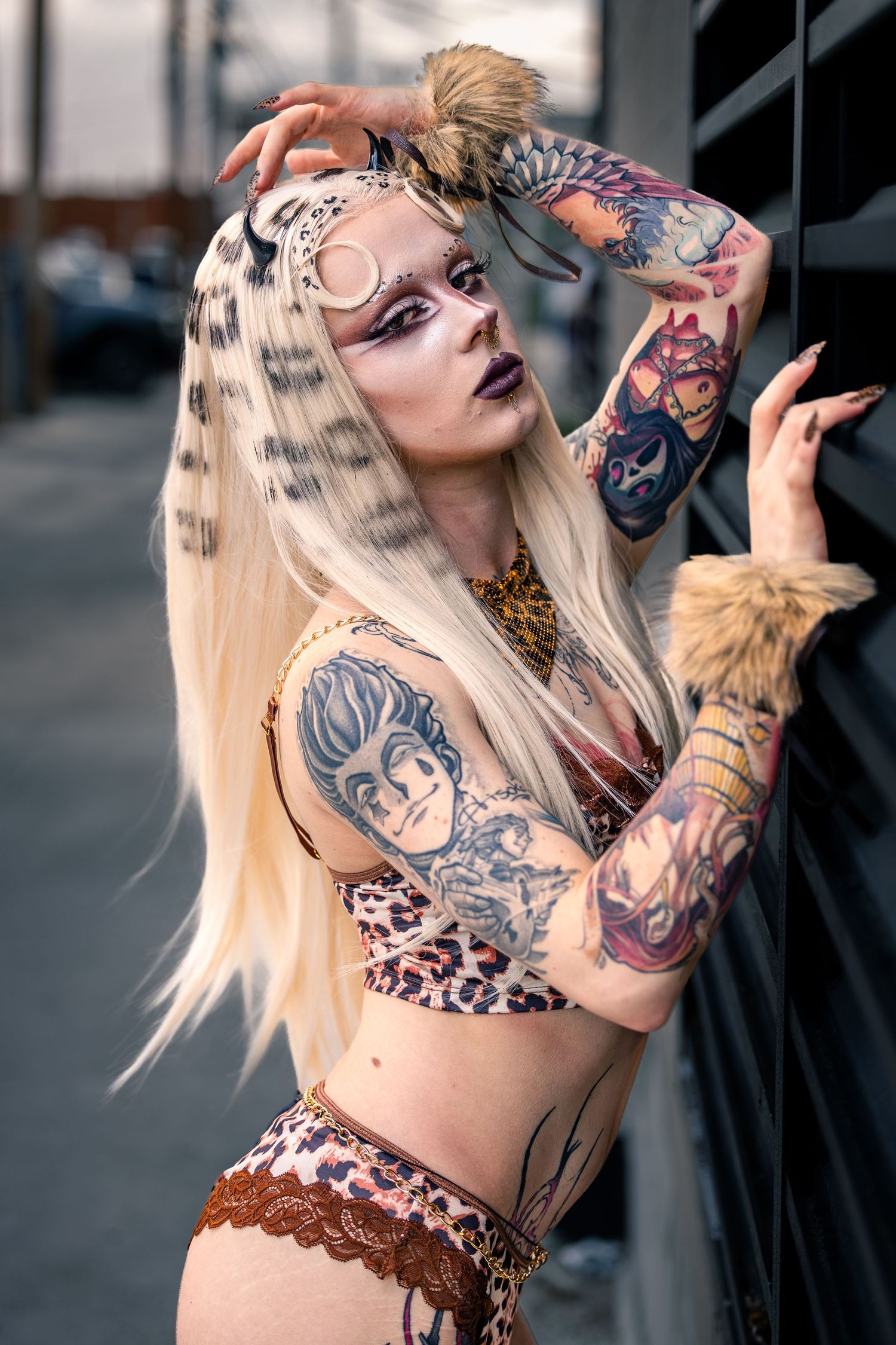 Heavily tattooed blonde woman in leopard print two-piece with dramatic makeup posing outdoors.