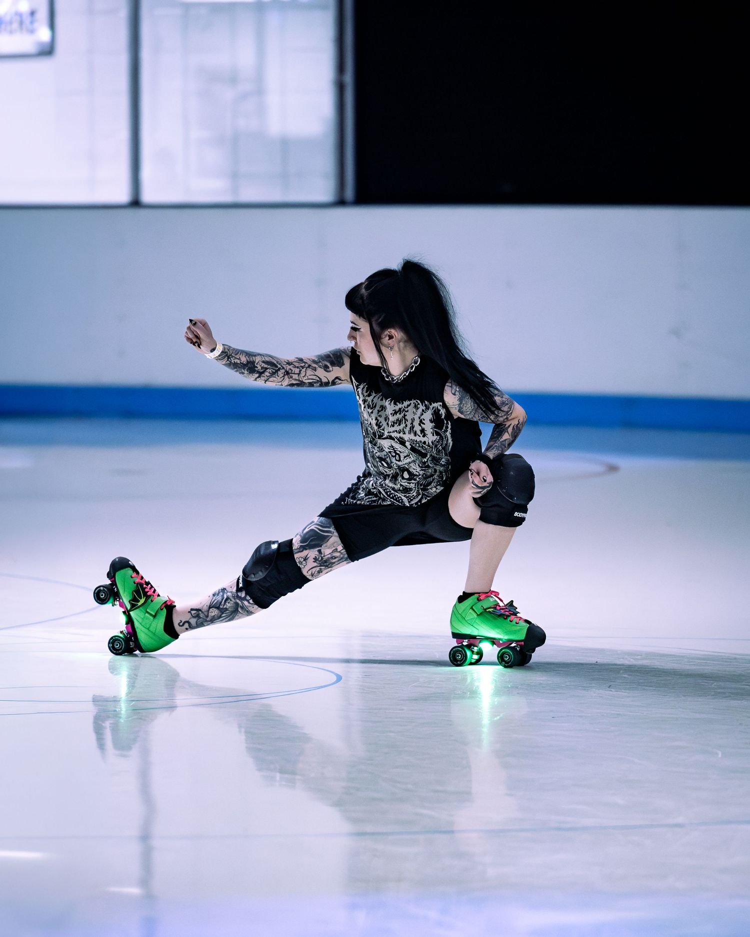 Tattooed woman performing a low lunge on green quad roller skates on an indoor skating rink.