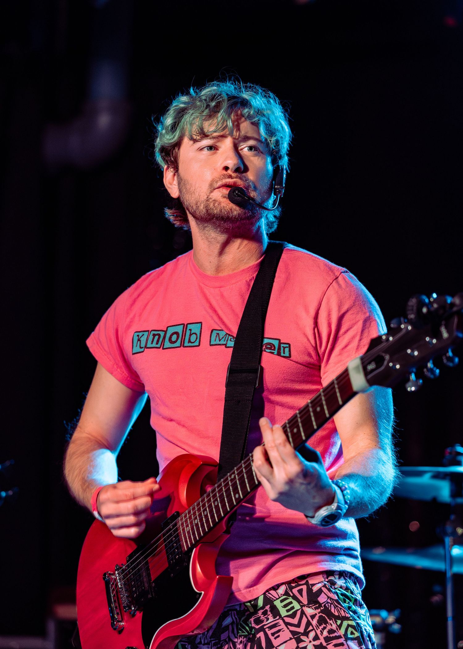 Musician with blue hair plays red guitar on stage wearing pink t-shirt under colorful stage lighting.
