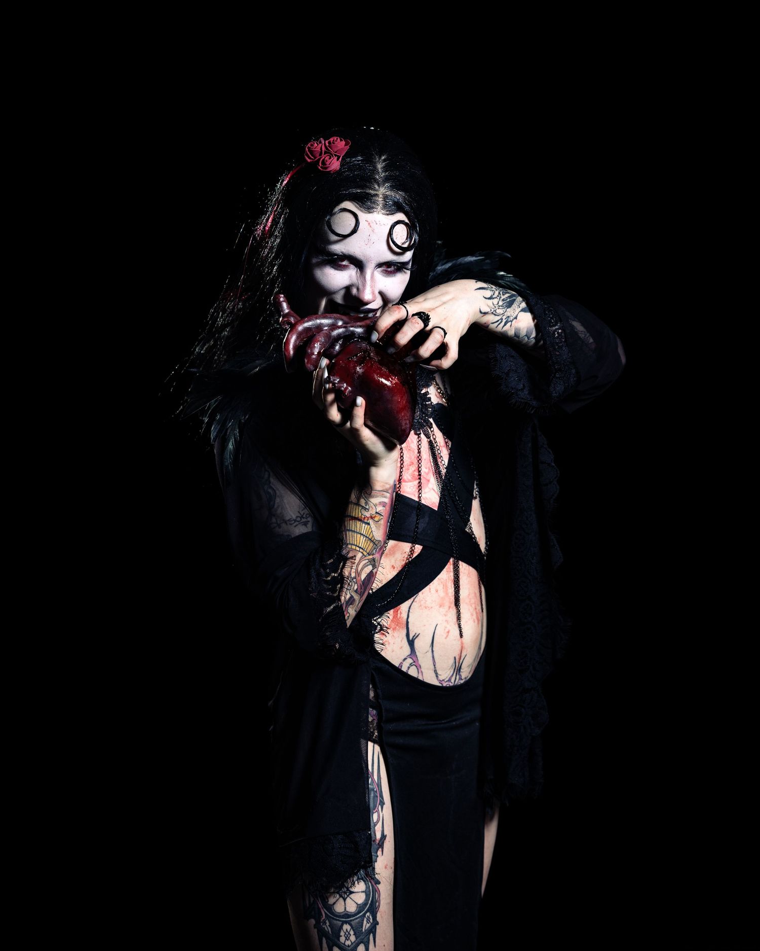 Person in horror costume with zombie makeup, black robe, tattoos, and bloody heart prop against dark background.