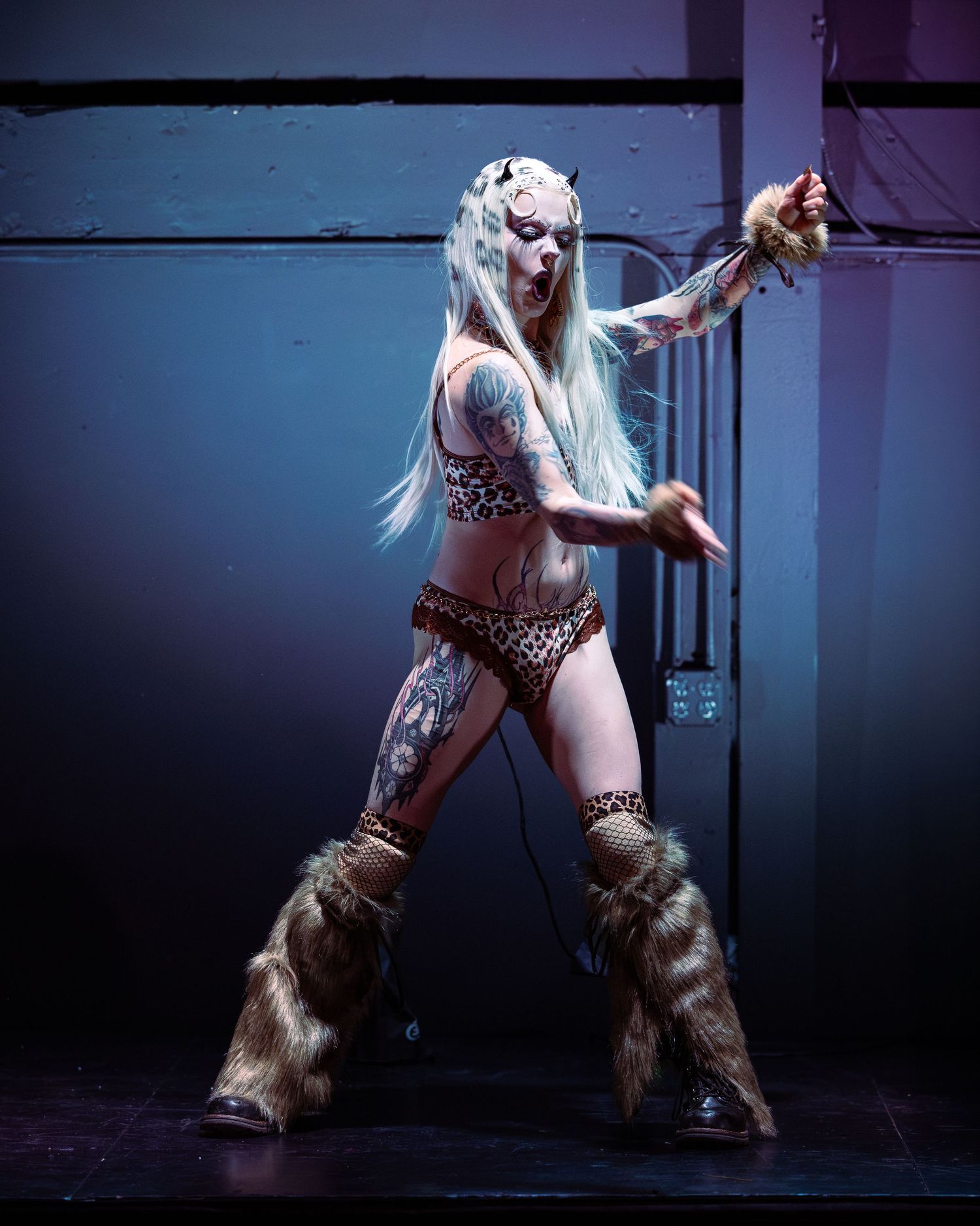 Tattooed performer in leopard print bikini and fur boots poses dramatically on stage with white hair.