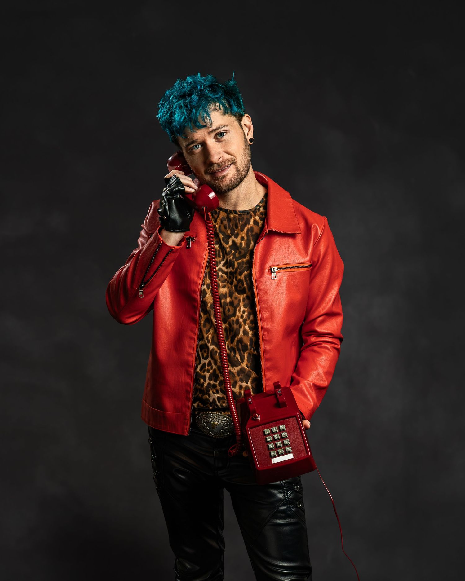 Teal-haired person in red leather jacket talking on a retro red telephone while wearing leopard print shirt.
