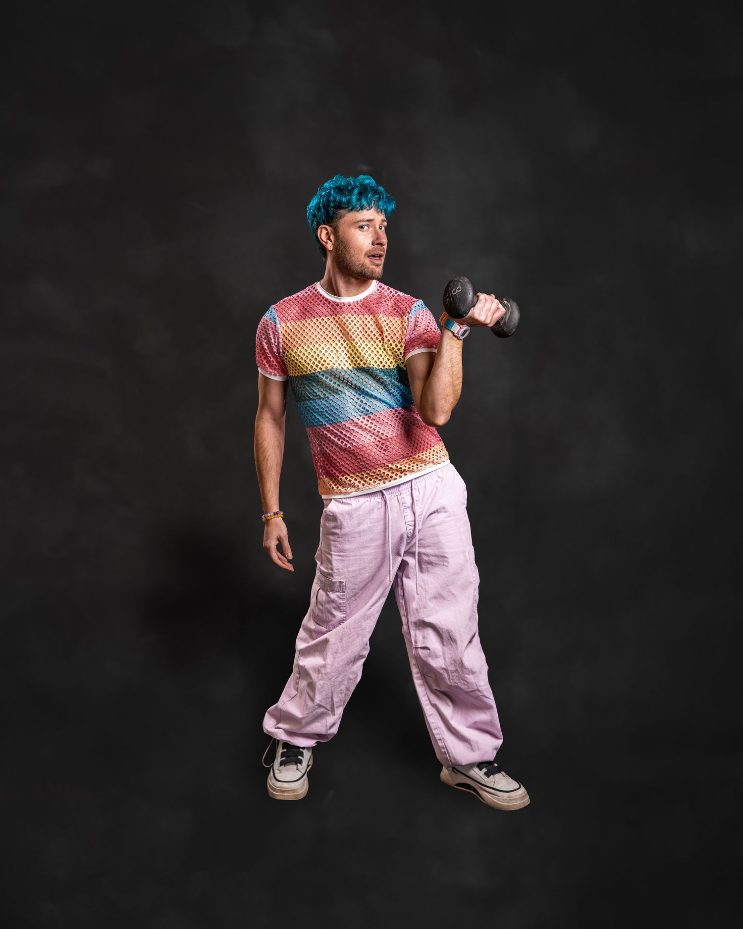 Man with blue hair in rainbow mesh shirt and pink pants curling a dumbbell against dark background.
