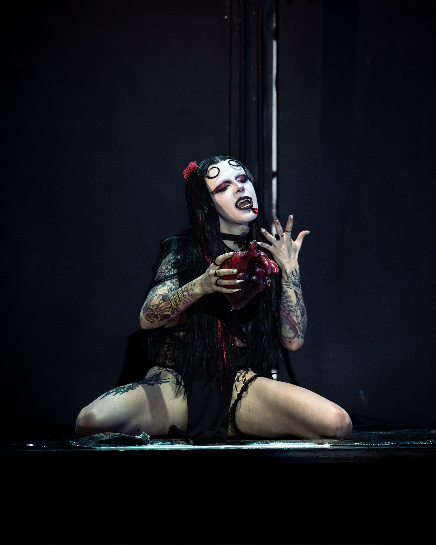 Gothic performer with white eyes makeup kneels on dark stage holding dripping red heart prop.
