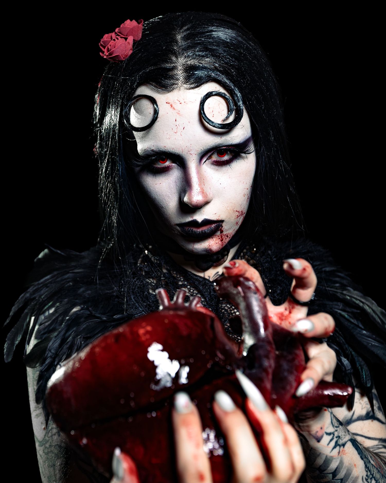 Close-up of gothic performer with white spiral eyes makeup holding bloody heart prop on dark background.