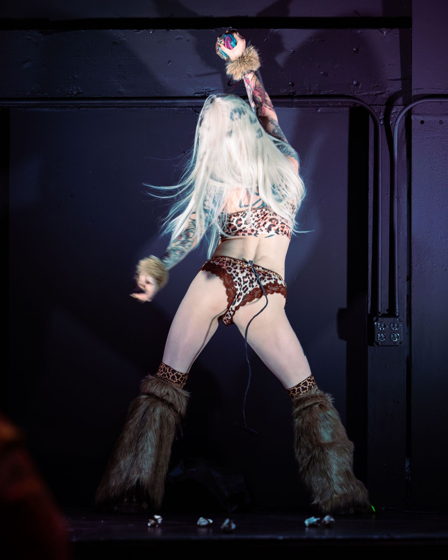 Performer in leopard print bikini and fur boots poses on stage with white hair and raised arm.