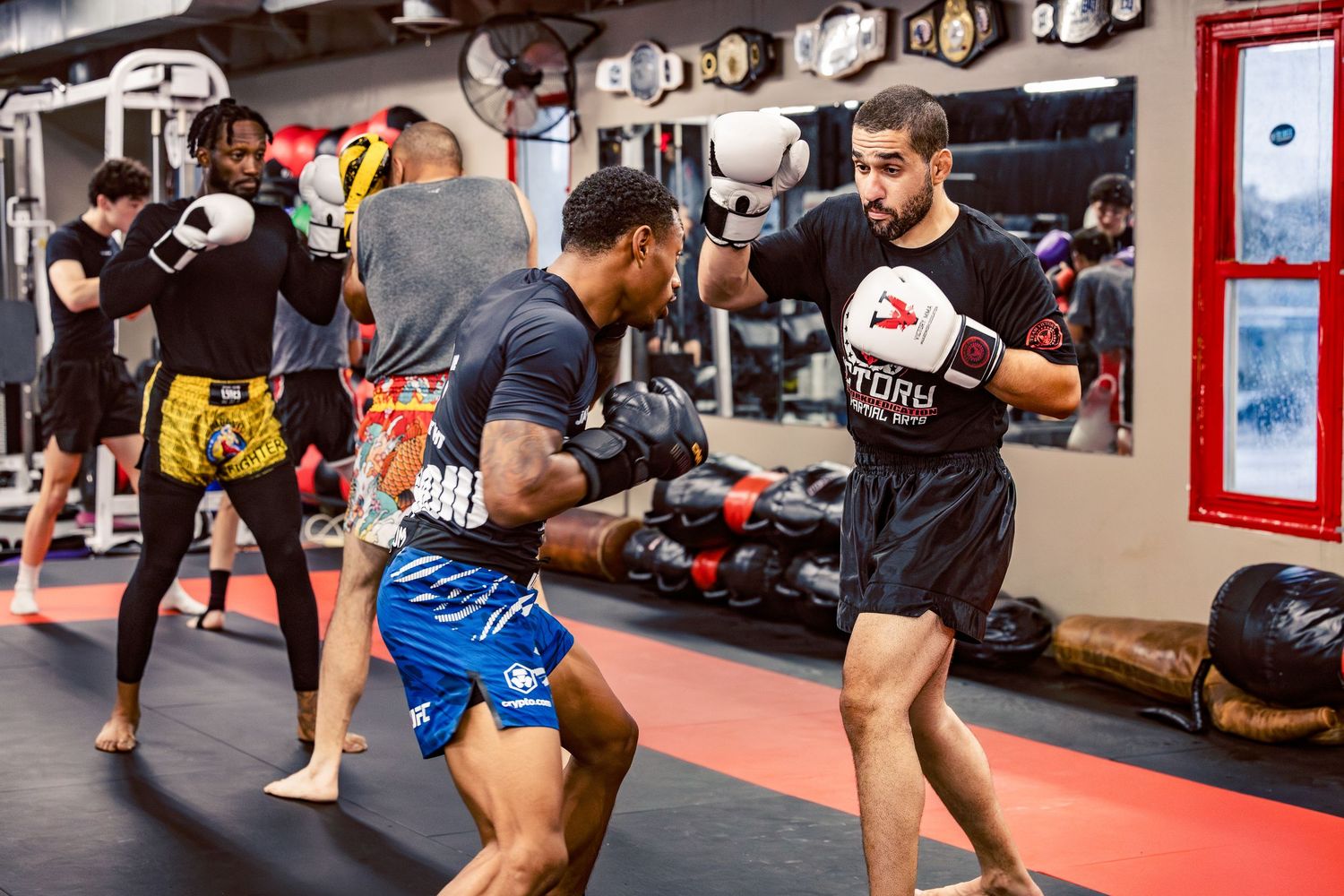 Mixed martial arts students practicing combat moves in a gym with championship belts on display.