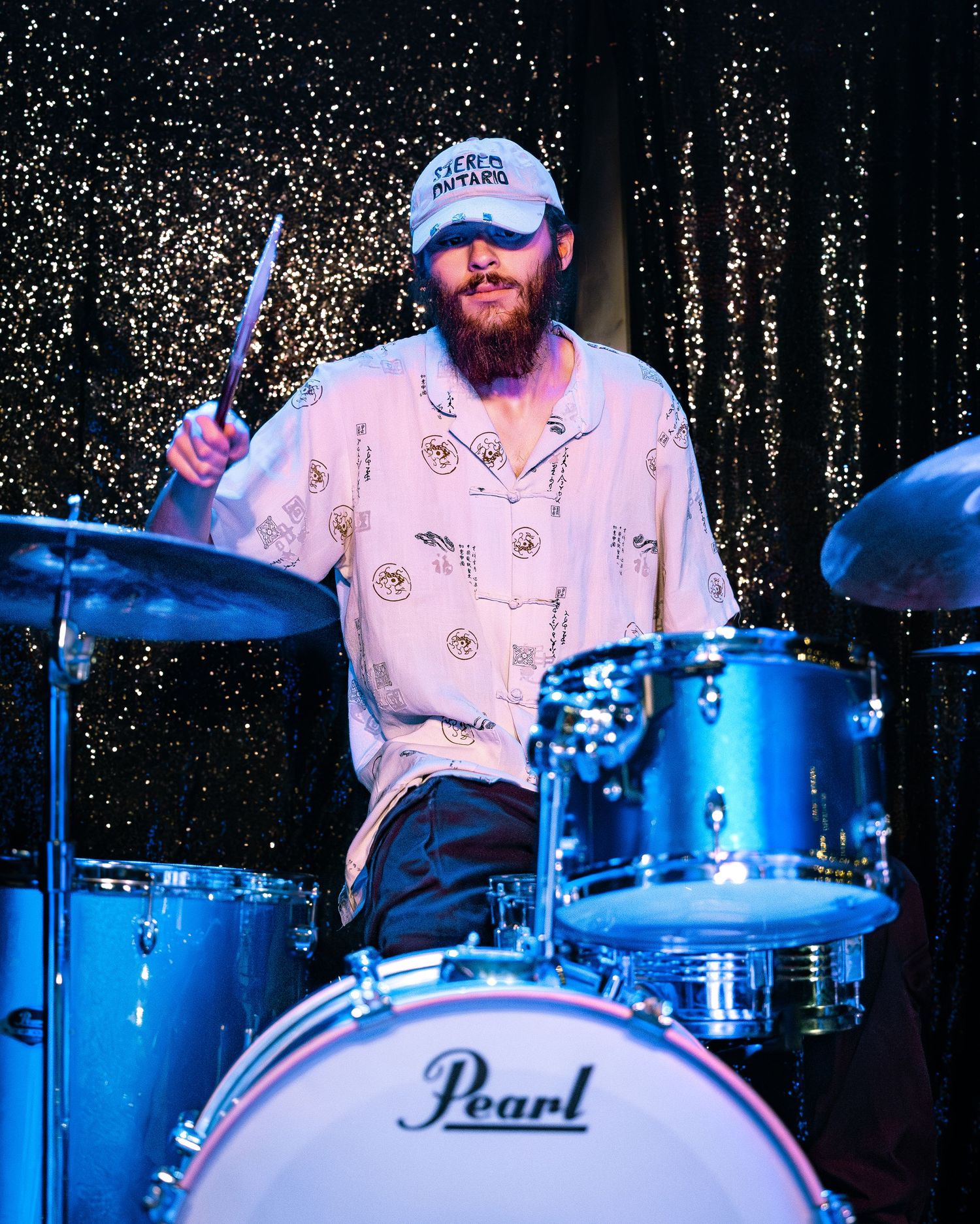Bearded drummer in white patterned shirt playing Pearl drum set at live music venue.