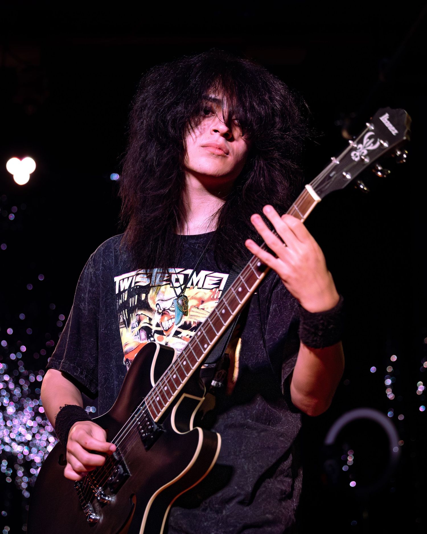 Young guitarist with voluminous dark hair playing a black electric guitar on a lit stage.