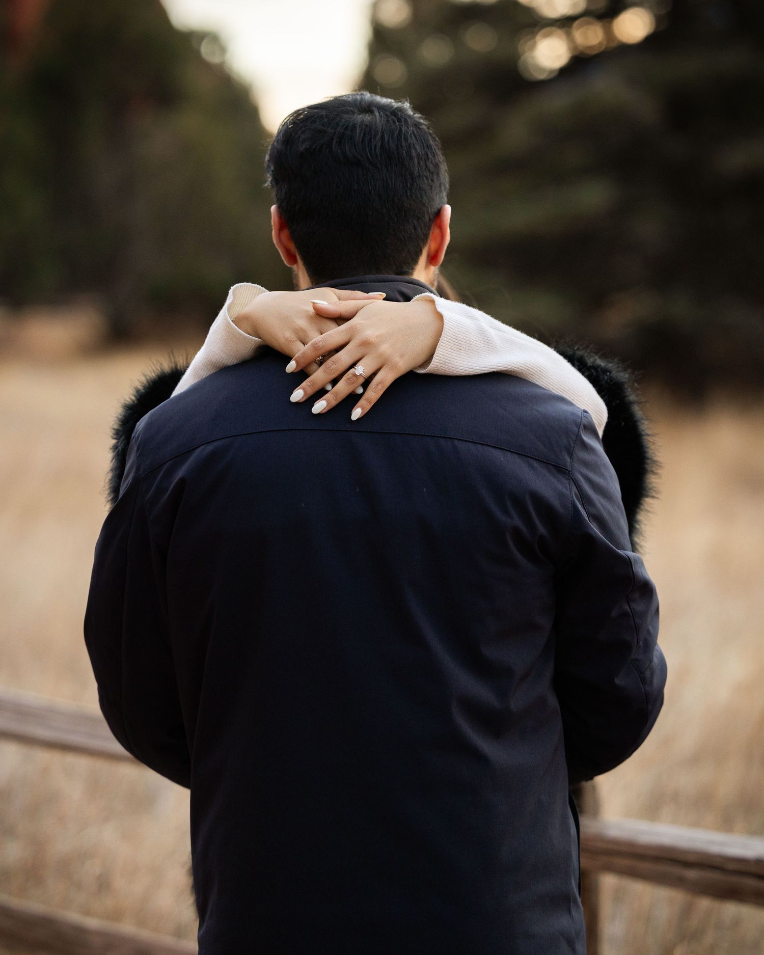 Person in black sweater seen from behind sitting on wooden bench in outdoor setting at sunset.