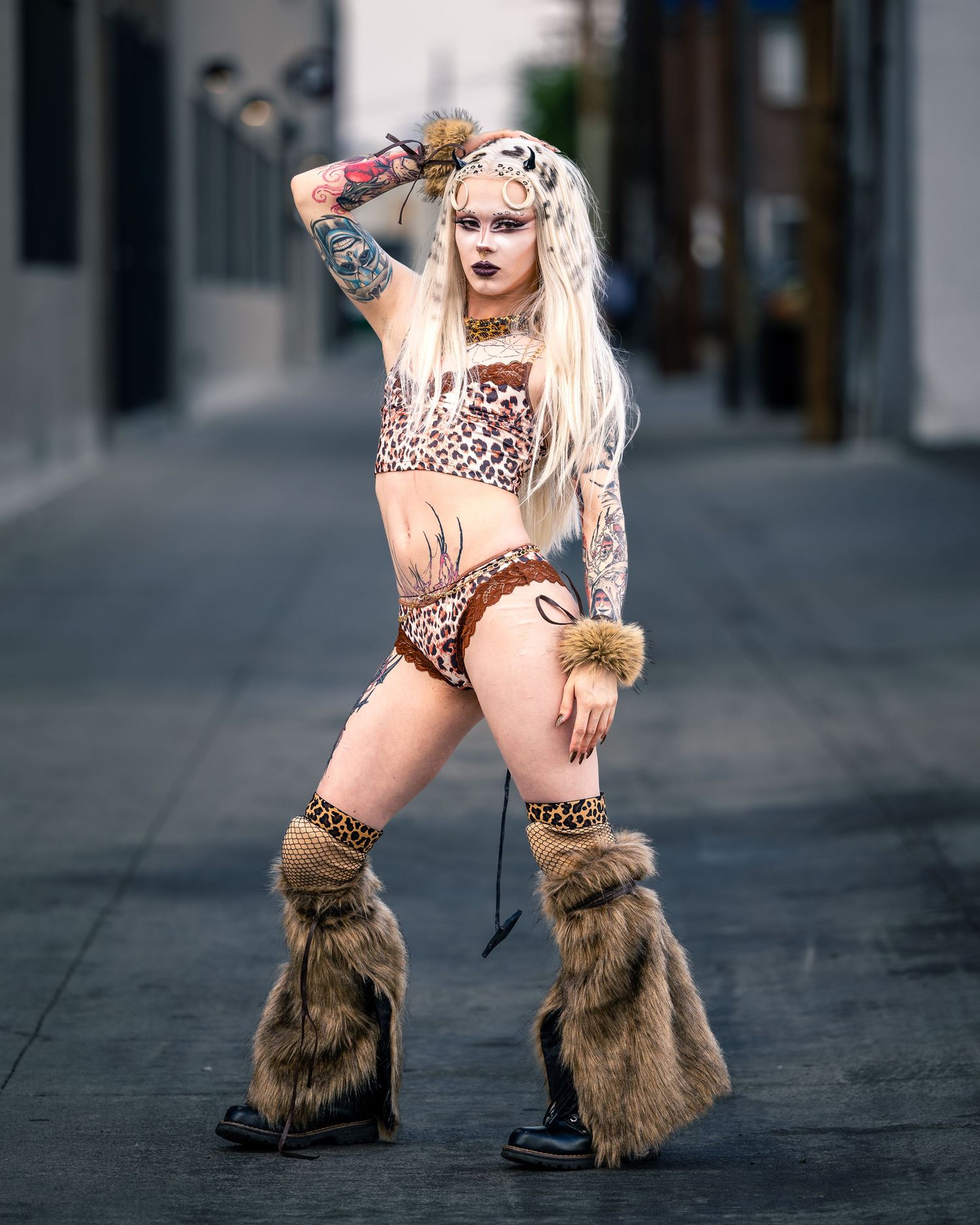 Tattooed woman in leopard print costume with fur leg warmers poses in urban alley.