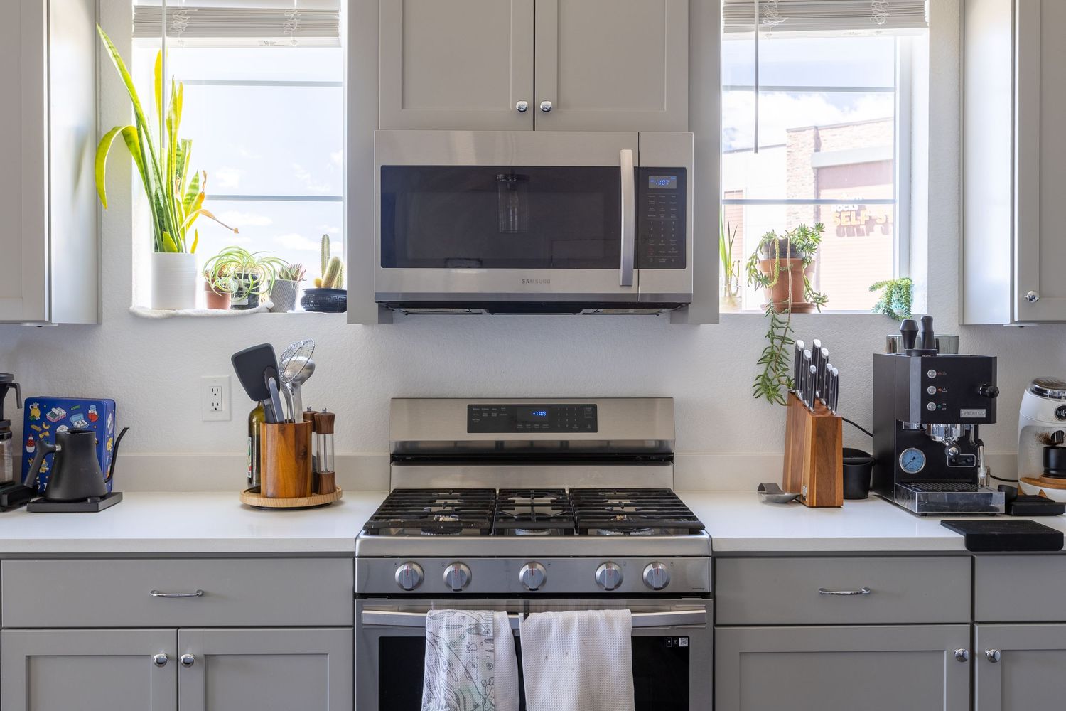 Modern kitchen with gray cabinets, stainless steel gas range, over-range microwave, and white quartz countertops near bright windows.