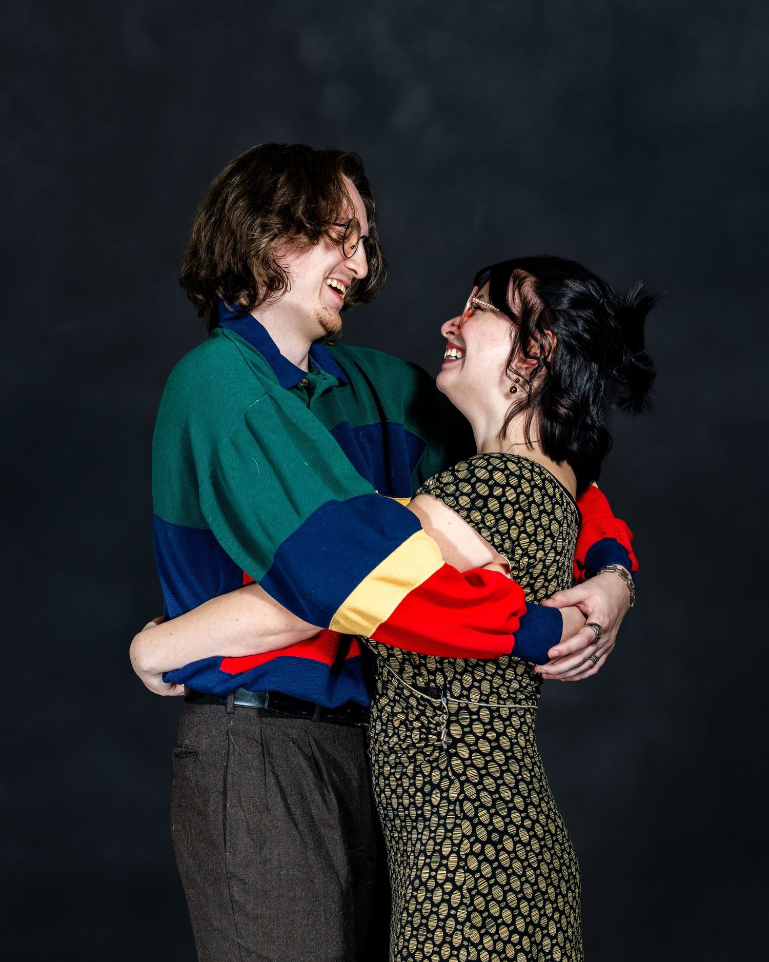 A joyful couple embraces and laughs together wearing colorful clothing against a dark studio background.