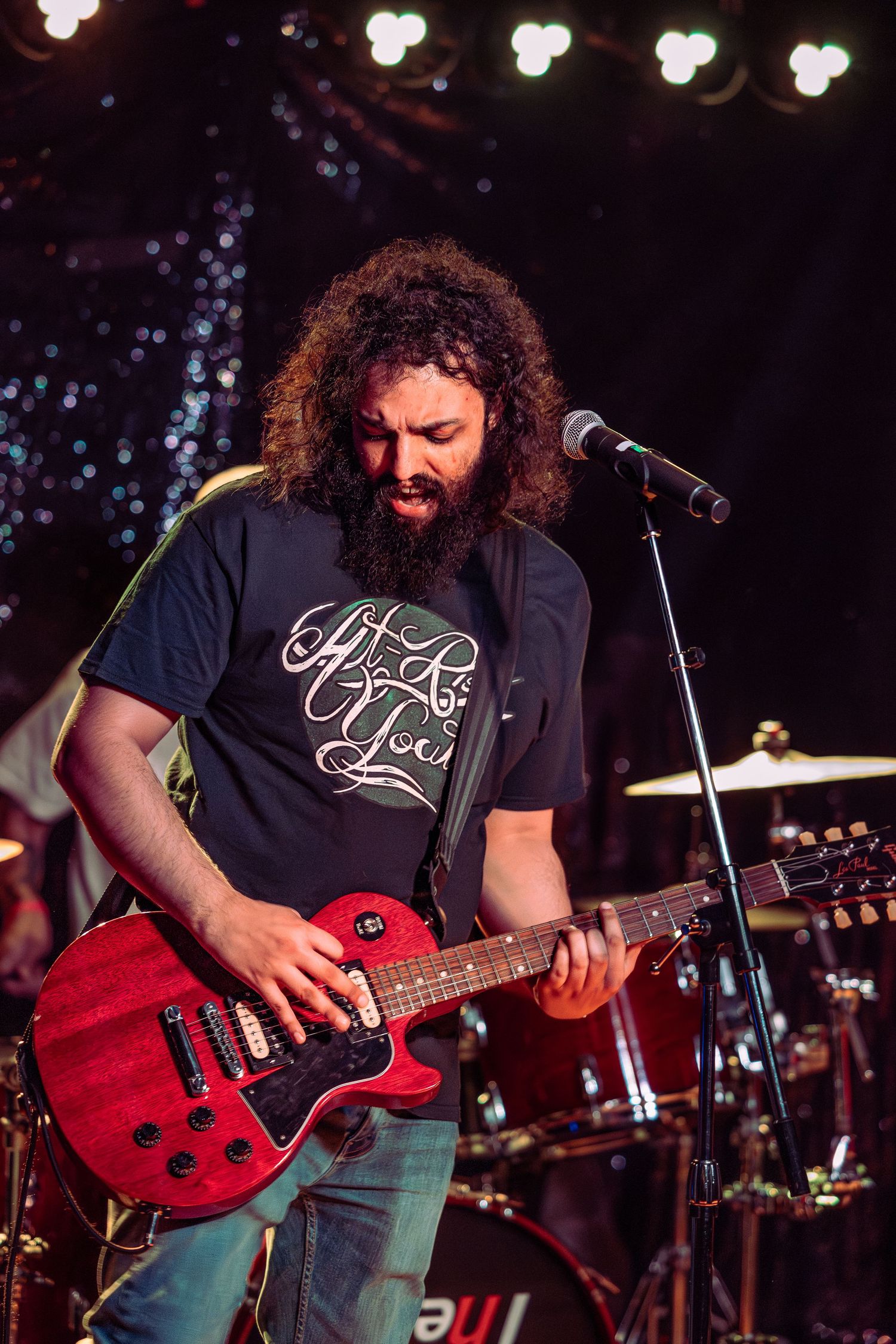 Bearded musician playing a red electric guitar on stage under colorful lights.