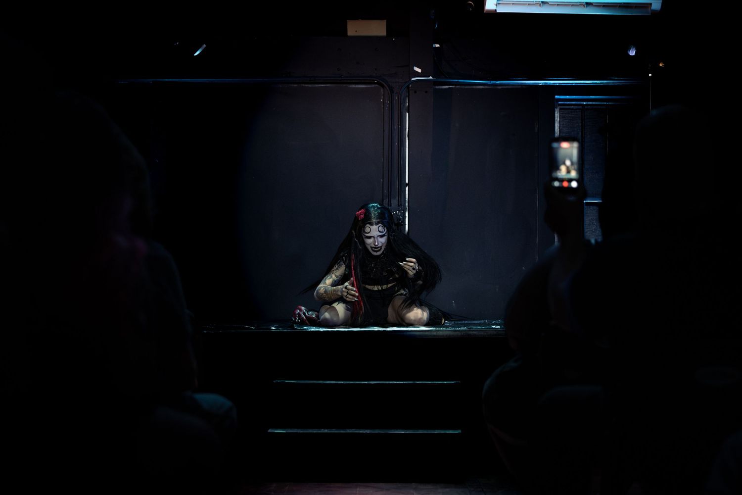 A performer in dramatic makeup sits illuminated on a dark stage, framed by silhouetted audience members.