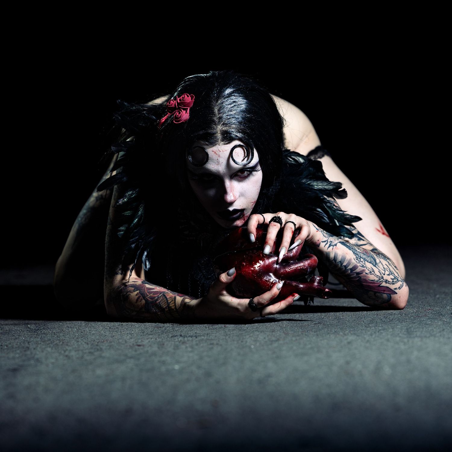 Gothic tattooed woman in dark makeup with black feathers lying on floor holding red object in dramatic lighting.