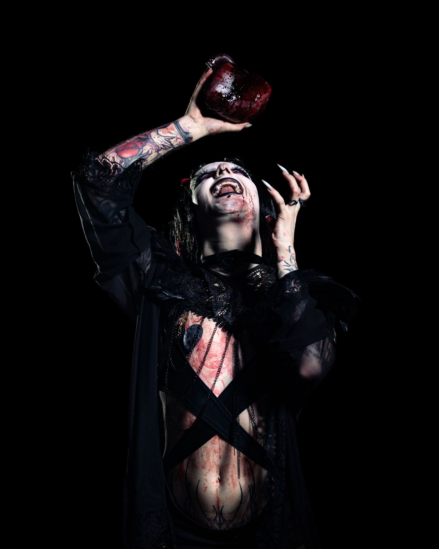 Gothic performer in dark makeup holding a prop heart, wearing black costume with tattoos on dark background.