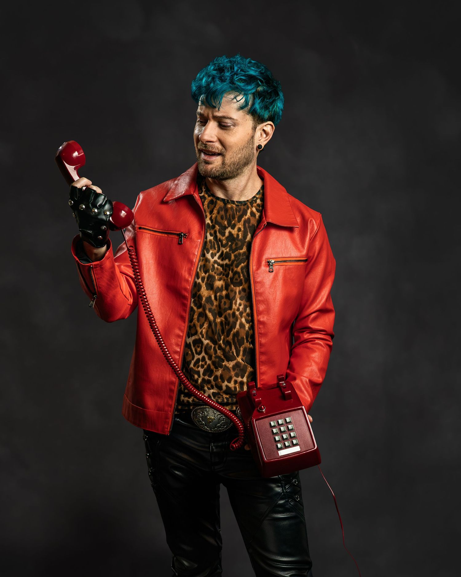 Man with teal hair in red leather jacket holds vintage red phone receiver, wearing leopard print shirt and black pants.