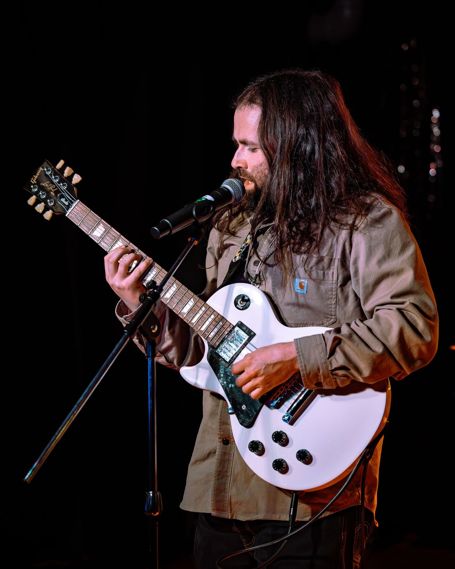 Smiling bearded guitarist with long hair playing a white Les Paul guitar on stage.